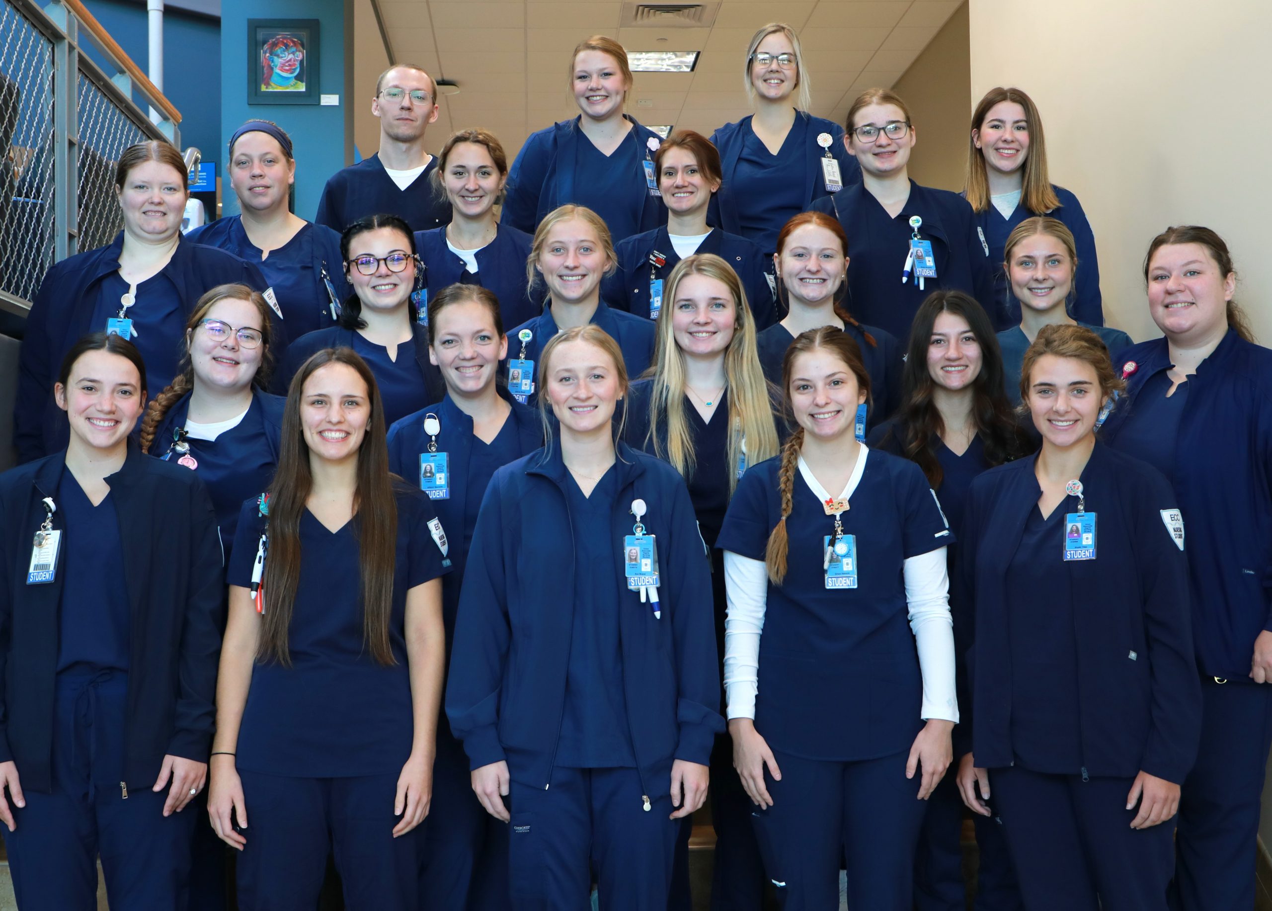 Group of 23 Nursing students dressed in blue clinical uniforms standing for a photo on a staircase