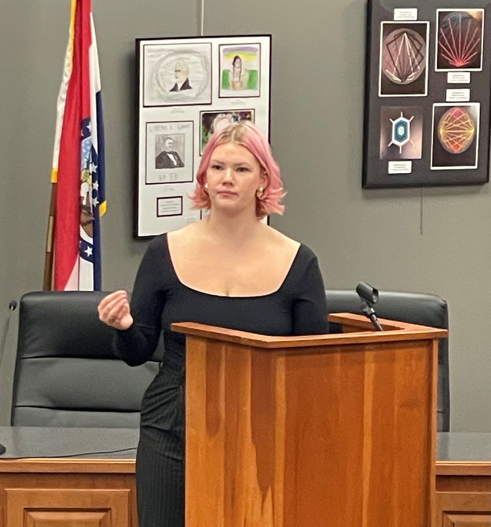 Lulu McGowan, PTK Heartland Region President, serving as the keynote speaker, stands at a wooden podium with pink hair, addressing an audience. She wears a black, square-necked top and appears focused on delivering her speech. The background includes framed artwork and a Missouri state flag.