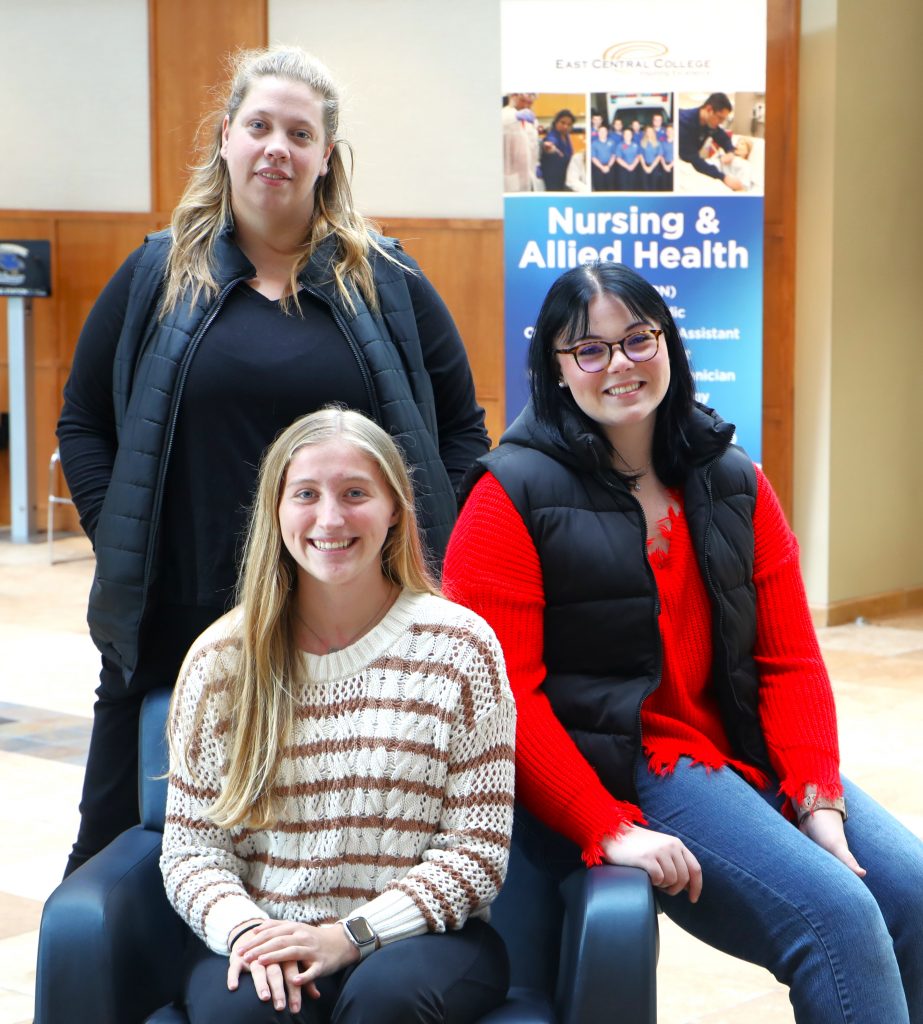 Three East Central College nursing students, Toni Sells, Olivia Reed , and Audrey Schroeder, pose together in front of a Nursing & Allied Health sign. These students, who recently provided life-saving assistance to a man injured in a car accident near the college campus, showcase their dedication and emergency response skills.