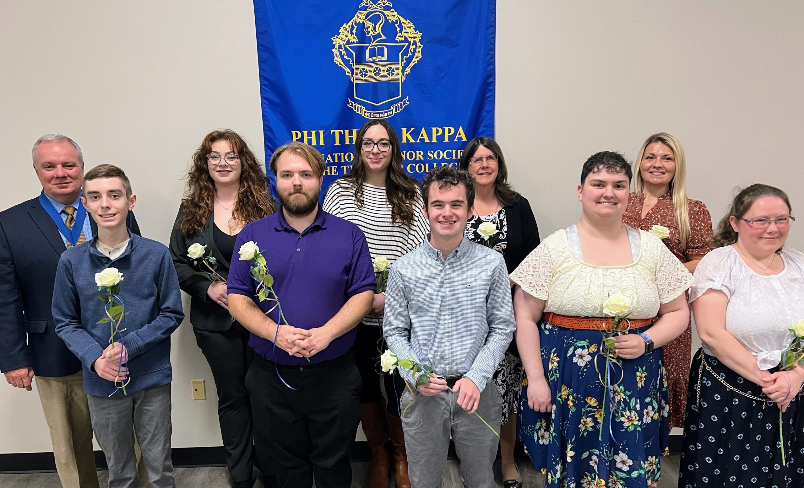 A group of Phi Theta Kappa (PTK) inductees and advisors from East Central College's Chi Delta Chapter stand together holding white roses. They are positioned in front of a blue PTK banner, smiling as they celebrate the Fall 2024 New Member Induction Ceremony at ECC Union.