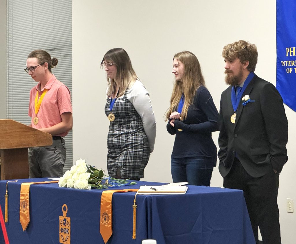 Four Phi Theta Kappa (PTK) members from East Central College’s Chi Delta Chapter stand at the front during the Fall 2024 Induction Ceremony. One member, wearing a pink shirt and gold PTK medallion, speaks at a podium, while the others stand beside a table draped with a blue PTK cloth, adorned with white roses and gold tassels.