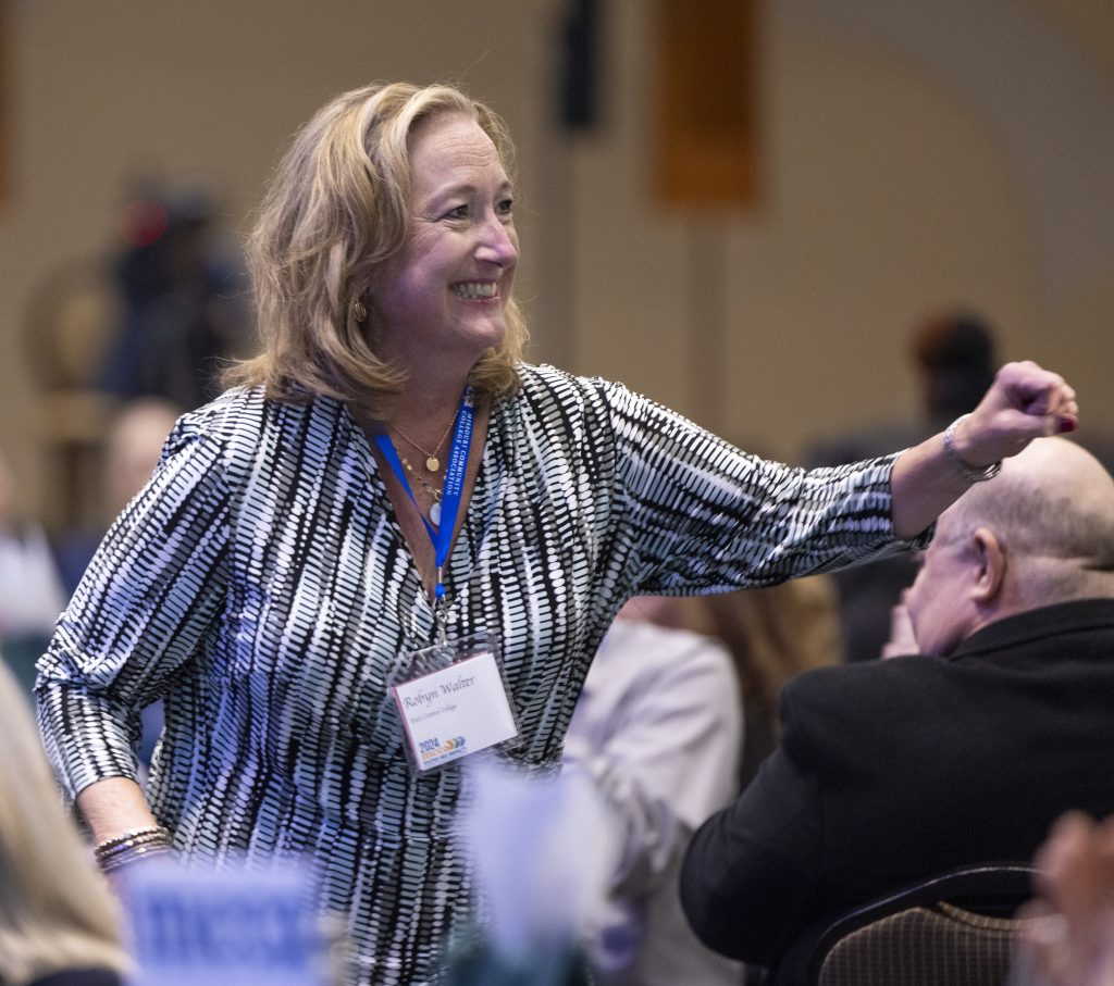 Dr. Robyn Walter, wearing a patterned blouse and a conference name badge, smiles and stands to acknowledge applause after being announced as the recipient of the Administrative Professional Award during the Missouri Community College Association (MCCA) convention. The background shows a room filled with attendees seated at tables.