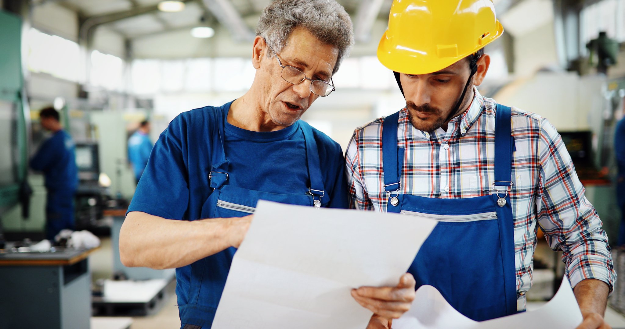An older male worker wearing glasses and blue overalls instructs a younger male worker in a hard hat and plaid shirt in a manufacturing setting. They are reviewing a technical document together, symbolizing training and skill development in the workplace.