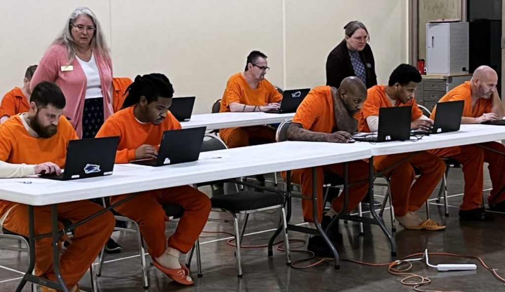 Detainees in orange jumpsuits sit at white tables in a classroom setting, guided by AEL instructors